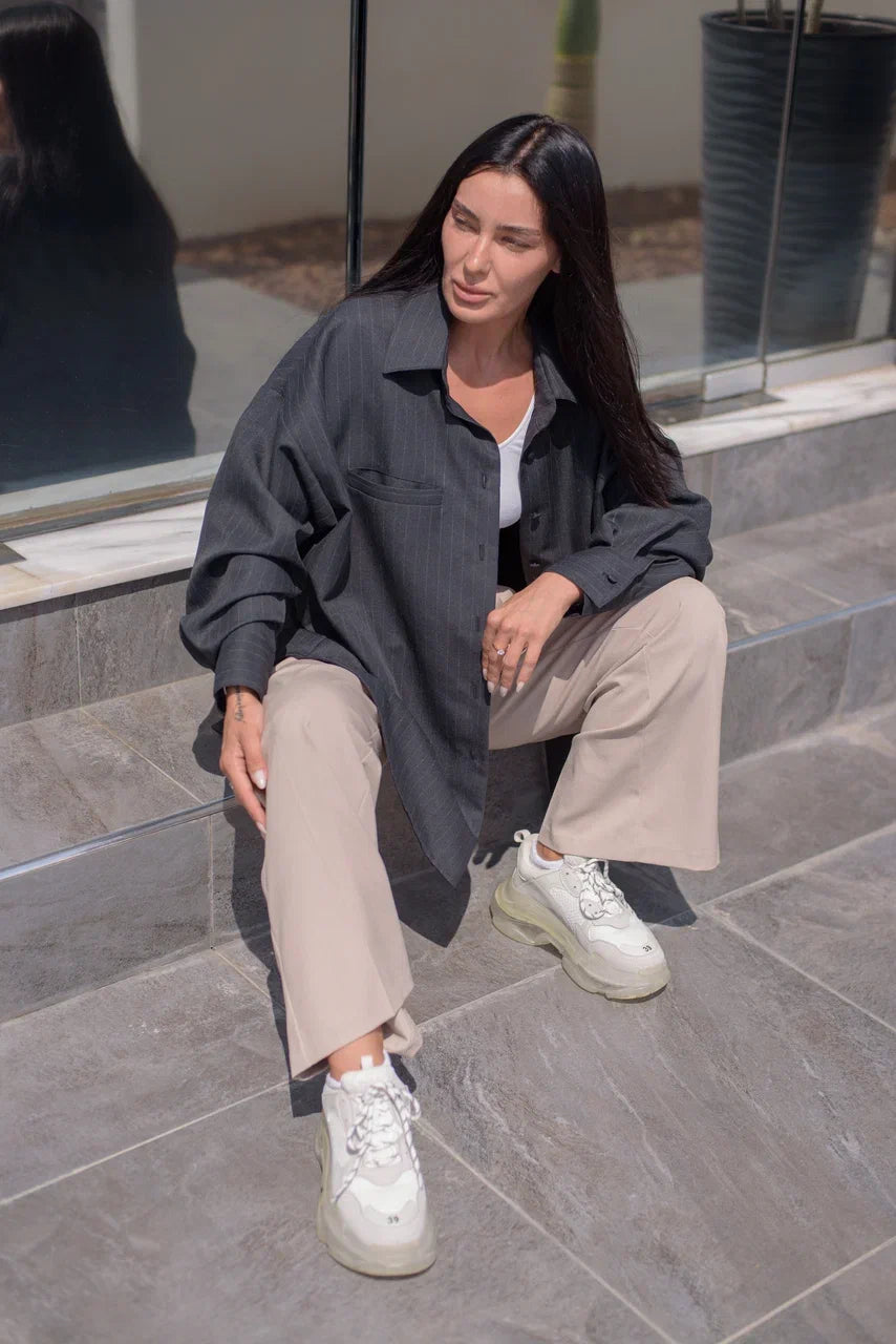 A woman sits on gray stone steps wearing a dark charcoal gray oversized button-down shirt, beige wide-leg trousers, and white chunky sneakers.