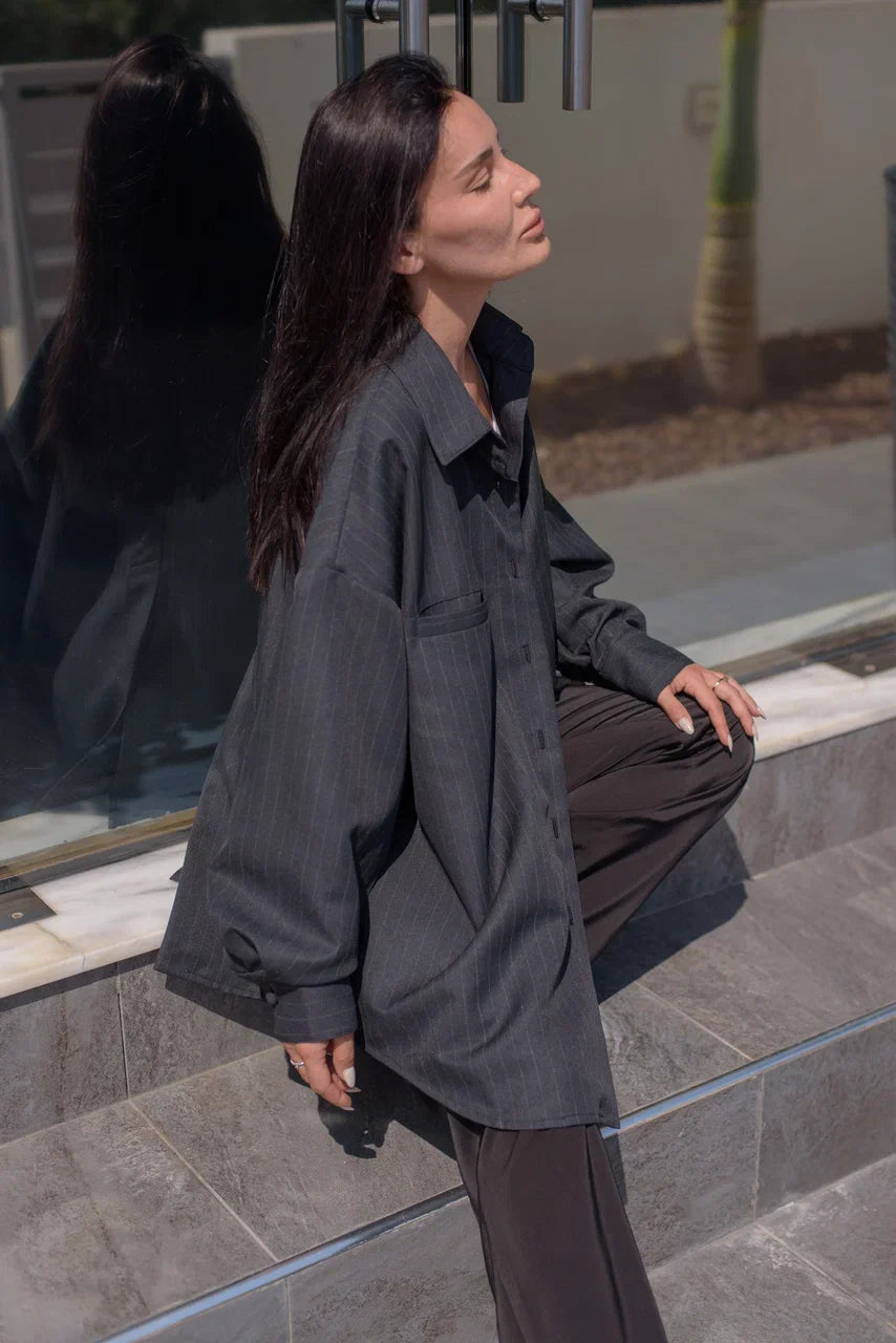A woman wearing an oversized dark gray button-up shirt and dark brown trousers, sitting on stone steps in an outdoor setting.