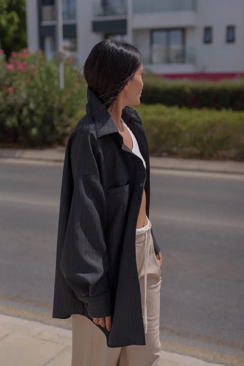 A woman standing outdoors on a sidewalk next to a road, wearing an oversized black pinstriped button-up shirt over a white top and beige trousers.