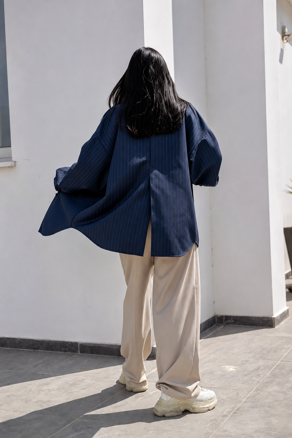 A person viewed from behind wearing an oversized navy blue pinstriped shirt jacket, beige wide-leg trousers, and chunky off-white sneakers, standing outdoors against a white wall.