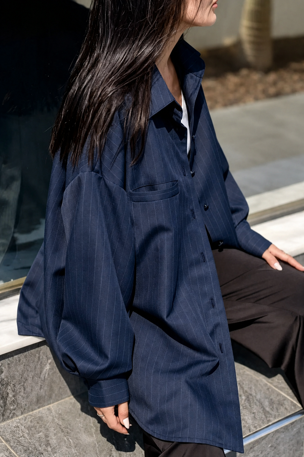A woman wearing an oversized navy blue pinstriped button-up shirt, styled with dark brown trousers, sitting on outdoor stone steps.